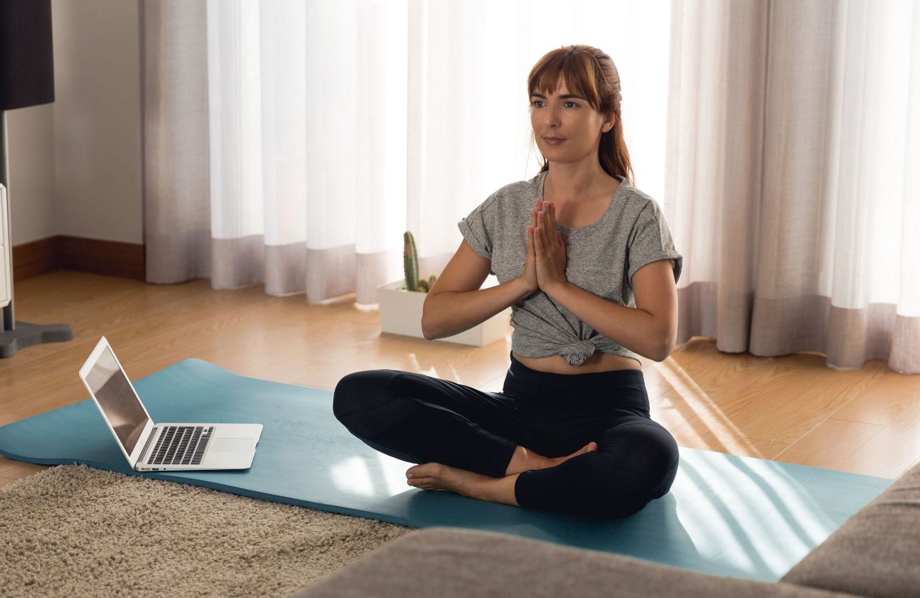 Frau sitzt im Schneidersitz auf ihrer Yogamatte und verfolgt einen Onlinekurs.