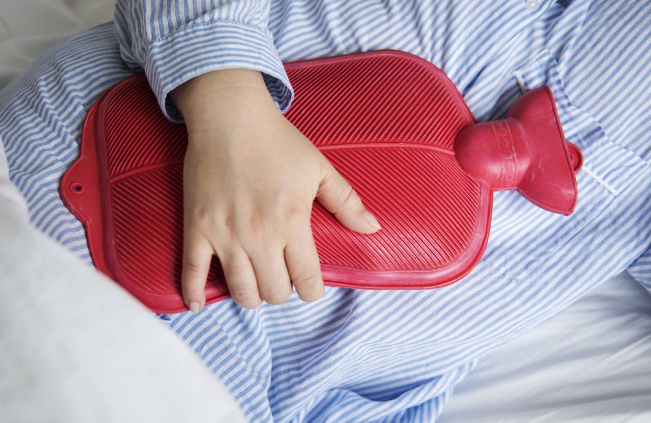 Person im gestreiften Pyjama hält sich wegen Bauchschmerzen eine rote Wärmflasche an den Bauch