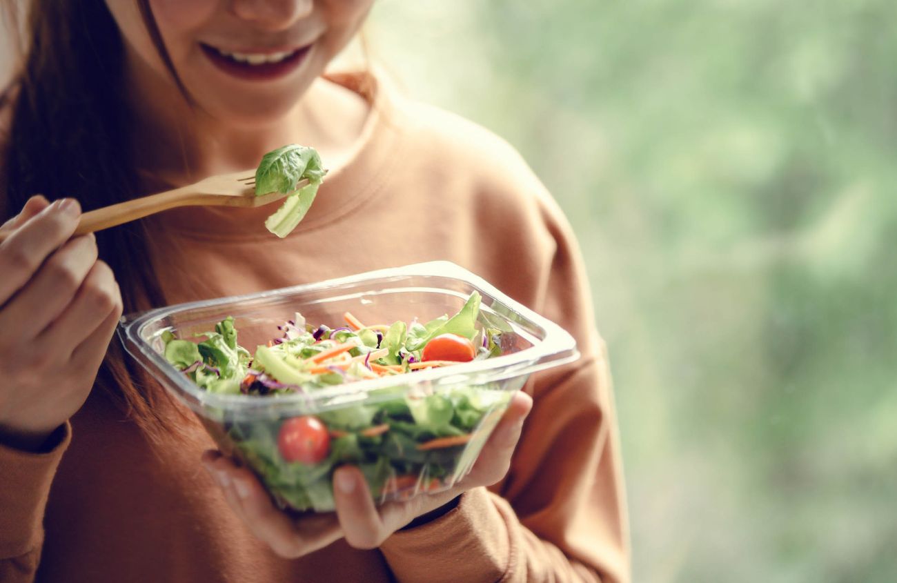 Frau isst einen gemischten Salat aus einer Plastikschale mit einer Holzgabel.