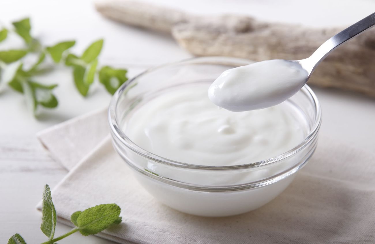 Eine Schale mit Joghurt steht auf einer Holzoberfläche. Ein Löffel schöpft daraus.