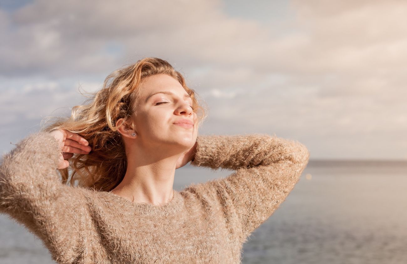 Frau bildet Vitamin D, in dem sie am Meer steht und den Kopf Richtung Sonne streckt