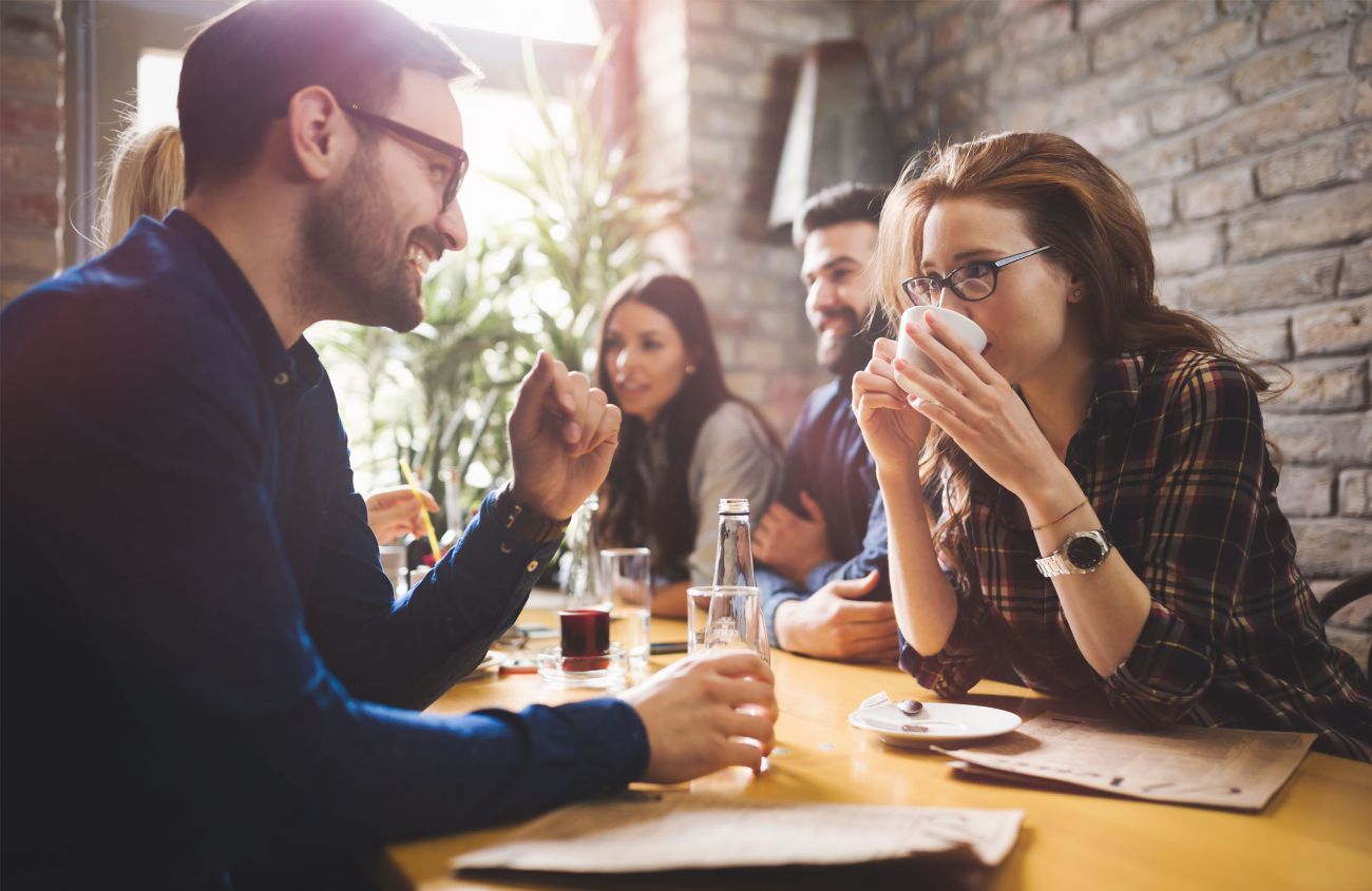 Eine Frau und ein Mann sitzen sich in einem Café gegenüber und unterhalten sich. Sie trinkt aus einer Tasse.