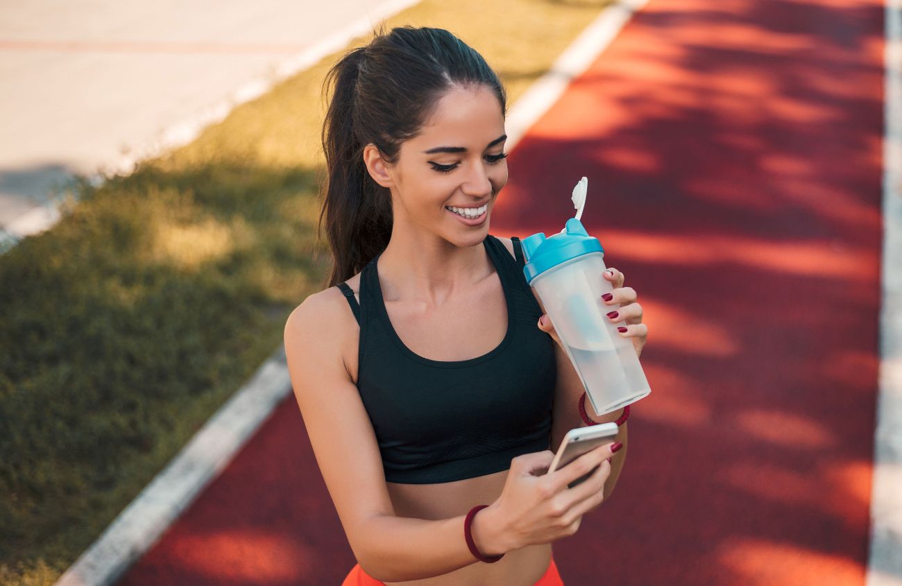Frau trinkt erleichtert Wasser und sieht auf ihr Handy: Sie hat Sport gemacht und fühlt sich wohl.