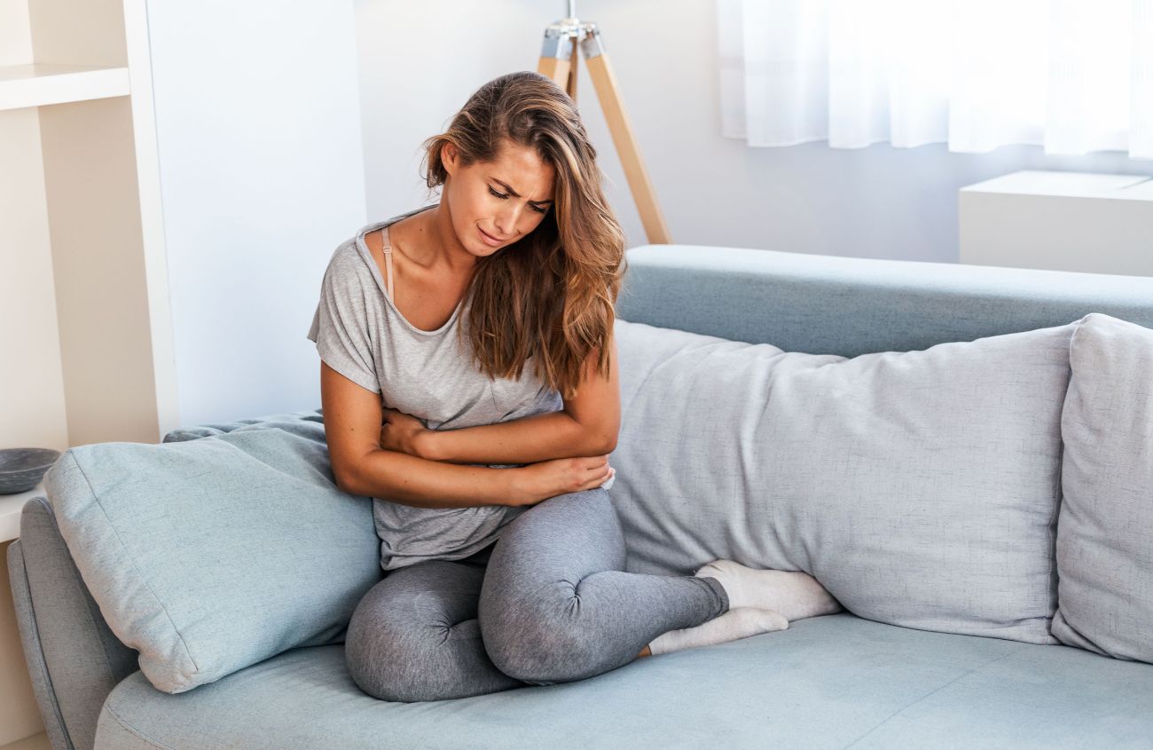 Frau sitzt mit schmerzverzerrtem Gesicht auf der Couch und hält sich den Bauch.