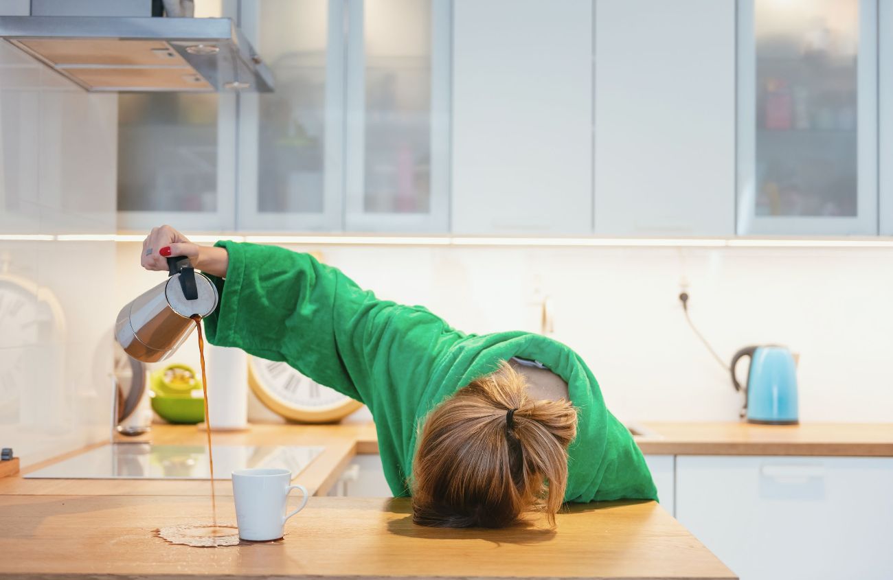 Frau im grünen Bademantel legt ihren Kopf auf den Tisch und verschüttet ihren Kaffee.