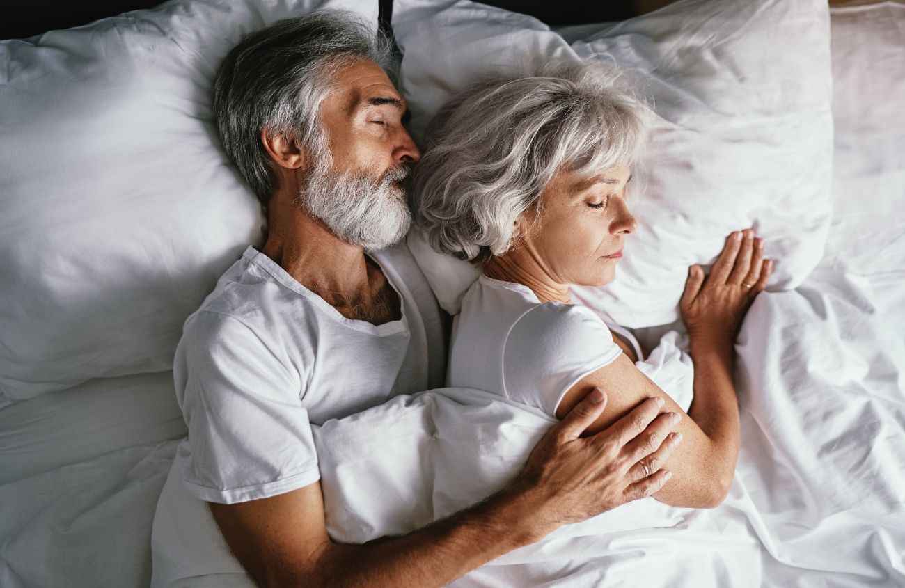 Ein älteres Paar liegt entspannt zusammen im Bett und schläft aneinandergekuschelt.