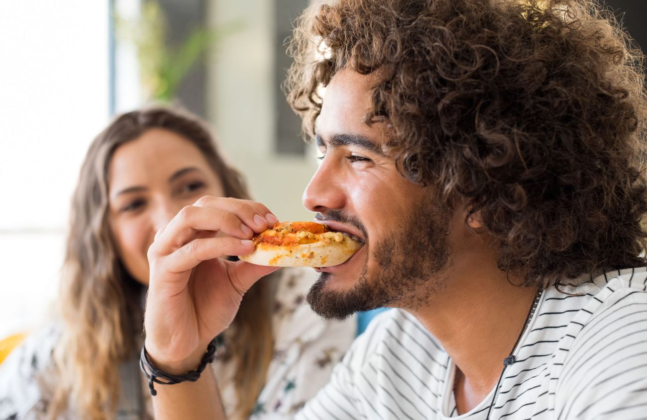 Ein junger Mann mit Bart und Locken isst eine Pizzaschnecke. Im Hintergrund sieht ihm eine Frau dabei zu.