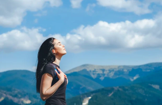 Frau streckt mit geschlossenen Augen den Kopf Richtung Himmel, im Hintergrund sind Berge zu sehen