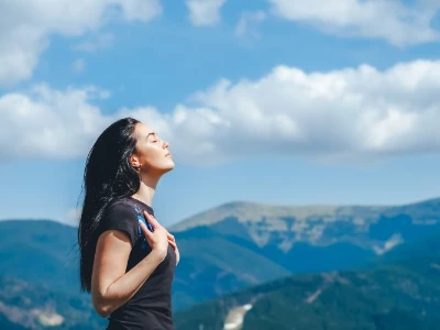 Frau streckt mit geschlossenen Augen den Kopf Richtung Himmel, im Hintergrund sind Berge zu sehen
