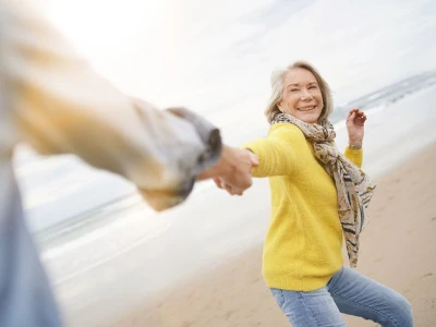 Eine ältere Frau in einer grell-gelben Jacke läuft am Strand und hält die Hand einer zweiten Person.