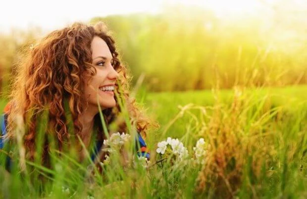 Frau mit langen lockigen Haaren liegt im Sonnenschein lächelnd in einer Wiese und schaut zur Seite