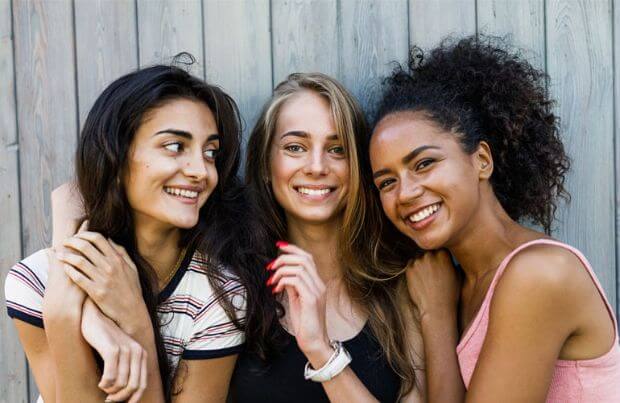 Drei junge, diverse Frauen stehen lächelnd vor einer Holzwand.