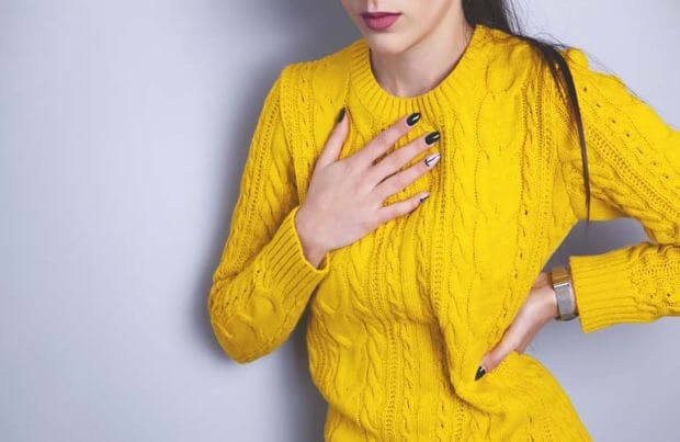 Frau im gelben Pullover fasst sich an die Brust: Sie leidet unter Sodbrennen.